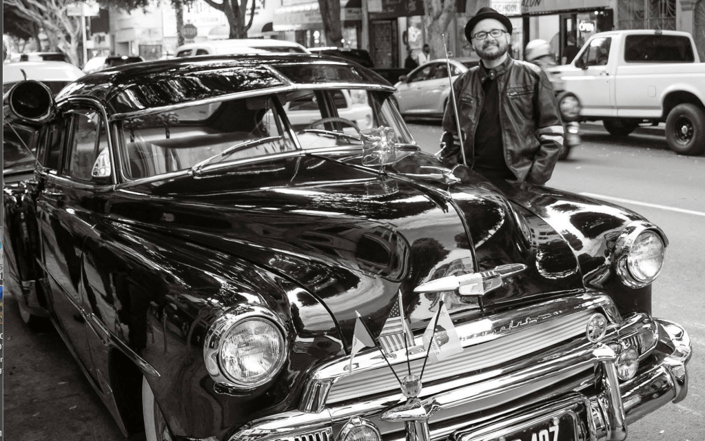 B&W photo. Drago is shown standing in front of a black bomba lowrider.